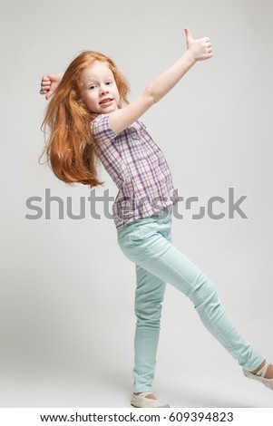 Vertical picture, isolated on grey, beautiful cute little caucasian redhead girl in plaid shirt, bright blue trousers and white boots