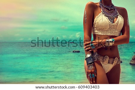Slim and tanned girl in trendy Boho style on the beach. Girl in a white knit swimsuit with silver jewelry . Bracelets and rings with turquoise stones.