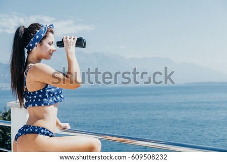 Beautiful glamour woman dressed in a swimsuit is standing in villa with hand binocular looking at sea. Copy space
