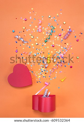 Red gift box in form of heart with various party confetti, streamers, noisemakers and decoration on a blue background. Colorful celebration concept.