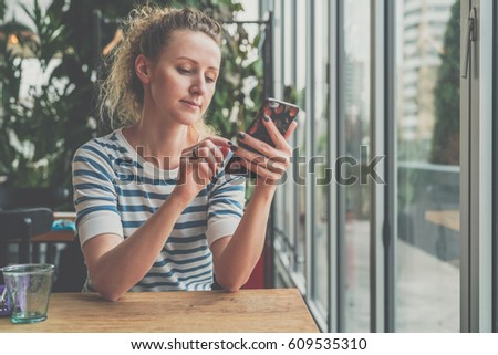 Young woman in striped blouse is sitting in cafe at wooden table near window and using smartphone.Girl shopping online,blogging,chatting,learning, checking email, browsing internet.Blurred background.