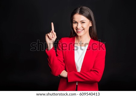 Young attractive businesswoman pointing up with finger and smiling at camera isolated on black