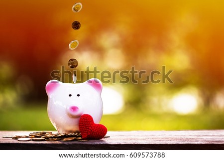 A piggy bank and dropping gold coins on the top and red heart put on the table in the public park, the saving for loved one and planned investment in the future concept.