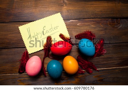 Red feathers with dyed, whole chicken eggs for the holiday with leaf and the inscription Happy Easter on wooden background.