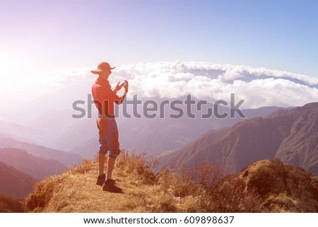 Person in sporty Clothing and Fedora Hat taking Photo of Mountain Scenery with curved Horizon and nice Cloud Cover of sunny Sky on Camera of Mobile Telephone