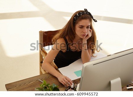 The upset female looks at the screen working at desk in front of computer in modern office. Tired designer or architect working at desk in design studio. Copy space for text