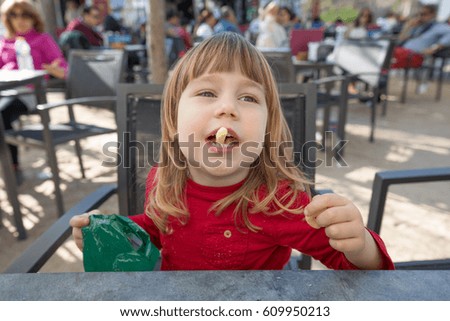 portrait of blonde three years old child face, with red shirt, eating cheese puff, with crumbs in mouth, sitting in terrace exterior bar cafe with grey table in park