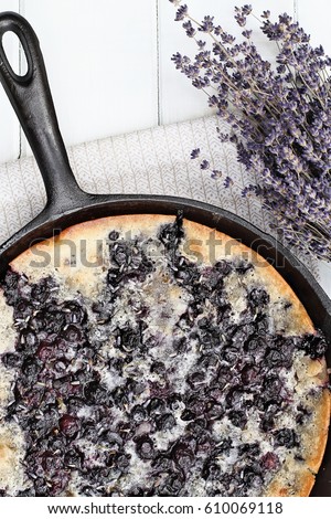 Above image of a blueberry lavender cobbler baked in a cast iron skillet over a white wood table top. Image shot from overhead. Perfect dessert for spring or summer.