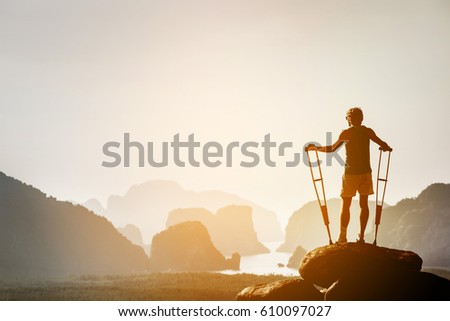 Disabled man with crutches stands on a big rock and looking at great view to islands and mountains. Space for text. Winner pose