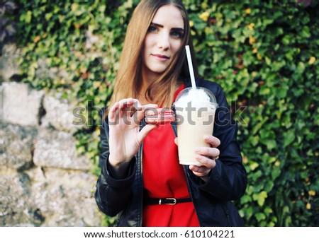 Outdoor closeup portrait of pretty stylish fashion girl having fun drinking chocolate milkshake with whipped cream and eating delicious macaroon. 