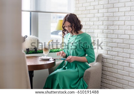 Beautiful girl in green dress sitting at the table and looks at the tablet in sunlight on the background of a white brick wall and windows