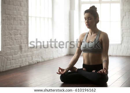 Young attractive yogi woman practicing yoga concept, sitting in Padmasana exercise, Lotus pose with mudra, working out wearing sportswear grey top and black pants, full length, loft studio background  Royalty-Free Stock Photo #610196162