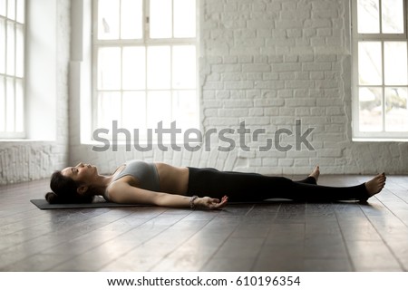 Young attractive yogi woman practicing yoga exercises concept, lying in Dead Body, Savasana, Corpse pose, resting after working out, wearing sportswear, full length, white loft studio background  Royalty-Free Stock Photo #610196354