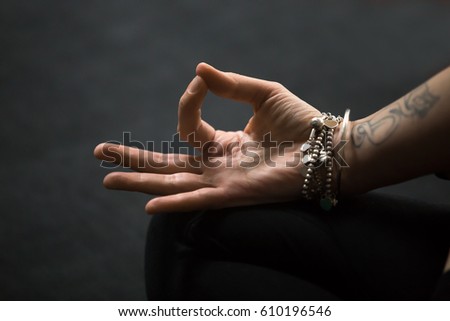 Close up of Dhyana (Gyan or Jnana) Mudra, spiritual hand gesture performed with yogi female fingers, tattoo and wrist bracelets. Woman meditating on black mat background. Meditation session concept  Royalty-Free Stock Photo #610196546