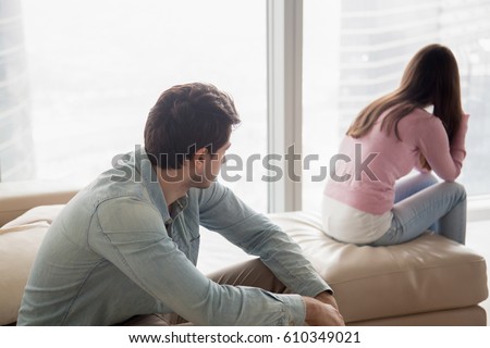Young couple sitting apart indoors after quarrel. Offended girl ignoring boyfriend, looking away, serious man thinking about problem in relationships, family conflict, misunderstanding between teens  Royalty-Free Stock Photo #610349021
