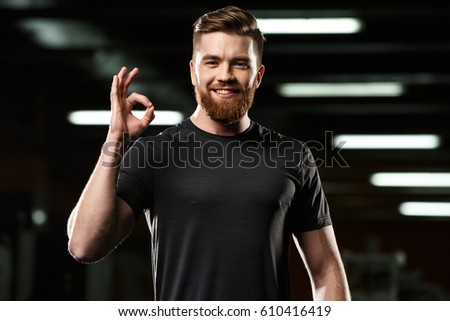 Image of cheerful sports man standing and posing in gym and looking at camera while showing okay gesture.