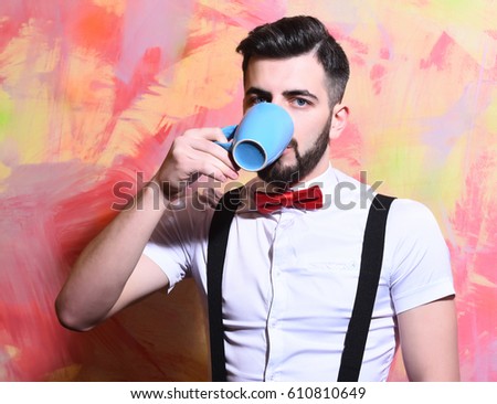 bearded man, long beard, brutal caucasian hipster with moustache in white shirt and suspenders with bow-tie drinking tea or coffee from blue cup with serious face on on colorful studio wall background