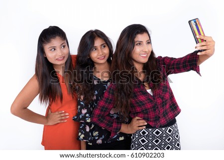 Studio shot of three happy young Persian woman friends smiling and posing while taking selfie picture with mobile phone togther against white background