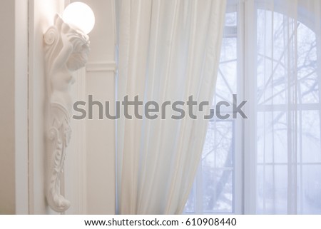 White caryatid holds a lighting bulb. Big window with curtain at the background