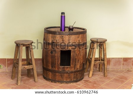 Barrel with perforation used as table. Two stools. Mate and violet thermo. Everything in the gallery of a house
