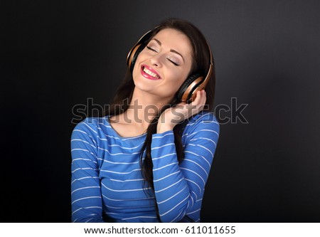 Beautiful toothy smiling young woman in blue clothing listening the music in wireless headphone with closed eyes on dark grey background
