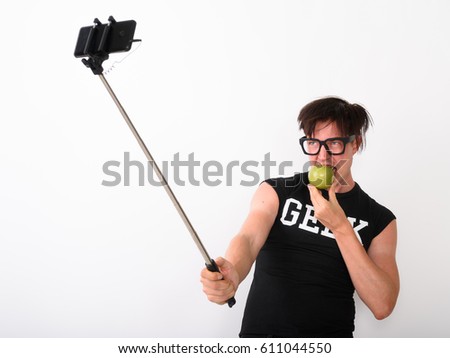 Studio shot of happy nerd man smiling while taking selfie picture with mobile phone on selfie stick and eating green apple and wearing shirt with geek text against white background