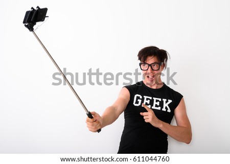 Studio shot of happy young nerd man smiling while taking selfie picture with mobile phone on selfie stick and pointing finger at phone while wearing shirt with geek text against white background