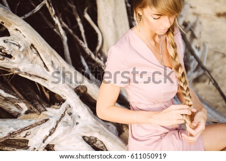 Young girl with long blond hair posing in a soft pink dress. She braids the braid from his hair. Concept of strong and healthy hair.  Romantic portrait. Pastel colours.Hipster toning. Soft focus.
