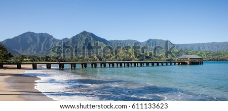 Famous Hanalei Pier in Kauai, Hawaii's North shore with beautiful ocean, beach, and green lush mountains in the background. Panorama Royalty-Free Stock Photo #611133623