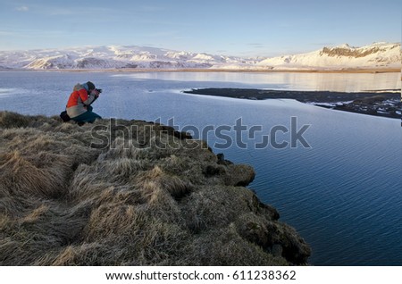 Photographer taking a photo on the grey grass with sea and mountains in background