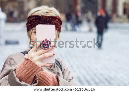 Face of a beautiful blonde woman coverd behind her pink phone in a cold winter in the historic district, when she takes pictures. She's wearing a winter jacket and ears are covered by headband.