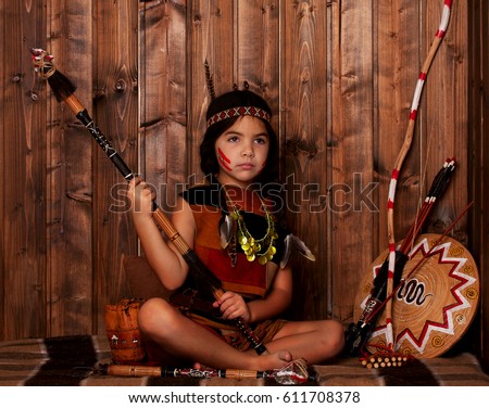 Little Girl disguised as an Indian holds in her hands on spear and laughs against the background of Indian entourage