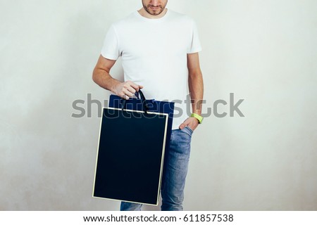 Young hipster man wearing white t-shirt and blue jeans, and holding black package with empty space for your logo or design, shopping bag with handles