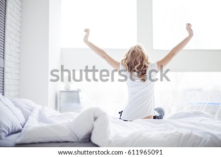 Blonde Woman sitting near the big white window while stretching on bed after waking up with sunrise at morning, back view. Royalty-Free Stock Photo #611965091