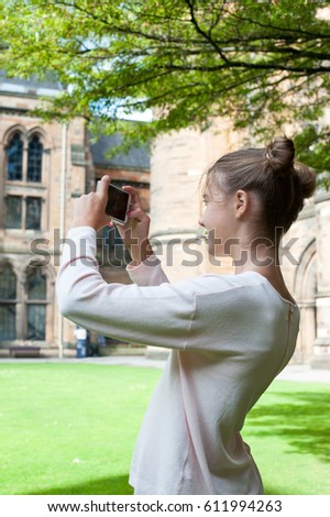 Young teenage girl sitting on the grass and taking picture with smatphone in Glasgow University garden. Vibrant colored summertime outdoors vertical image.