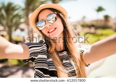 Cheerful young woman in hat and sunglasses taking selfie with mobile phone on summer resort palms background. 