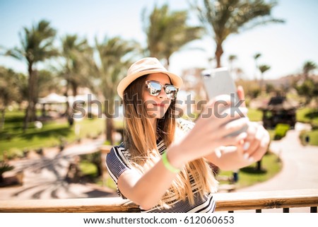 Cheerful young woman in hat taking selfie with mobile phone on summer resort