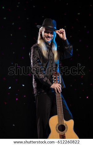 An elderly gray-haired male musician with long hair with a guitar in his hands is playing and posing on stage on a black background in a blue scenic light
