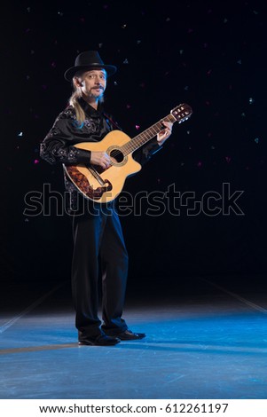 An elderly gray-haired male musician with long hair with a guitar in his hands is playing and posing on stage on a black background in a blue scenic light