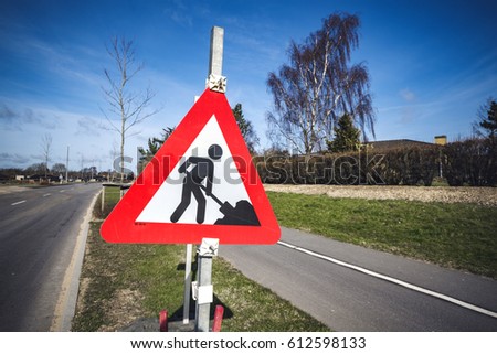 Road construction sign by a roadside in a city on a sunny day with blue sky