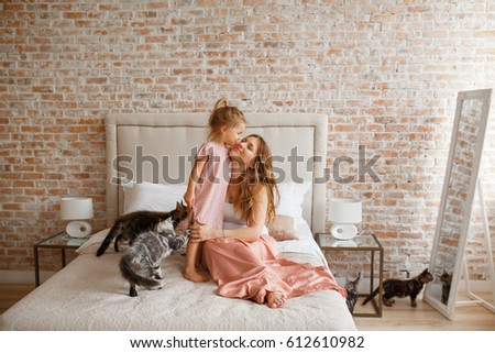 Mother and daughter having fun at home. Sweet little girl is hugging and kissing her beautiful young mom in cheek while sitting on bed at home, cats around