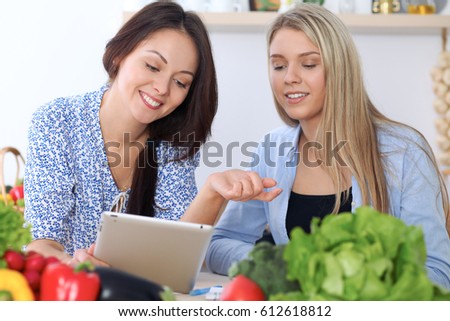 Two young happy women are making online shopping by tablet computer and credit card. Friends are going to cook in the kitchen, they searching internet for a new recipe