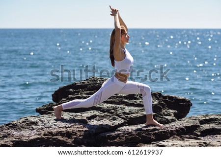 Young woman doing yoga in the beach. Female wearing sport clothes in warrior 1 figure with sea at the background.