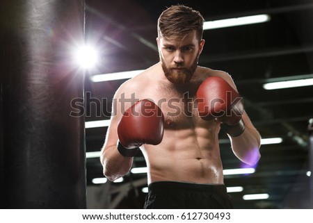 Picture of serious young strong sports man boxer posing in gym and looking at camera.