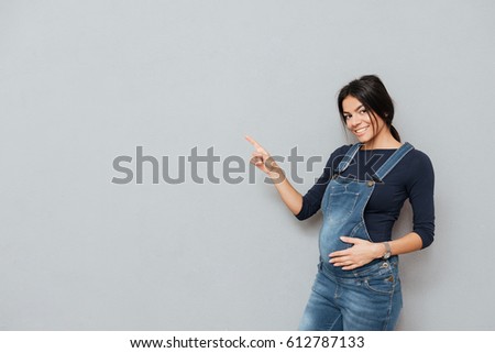 Picture of cheerful pregnant lady standing over grey background and pointing. Looking at camera.