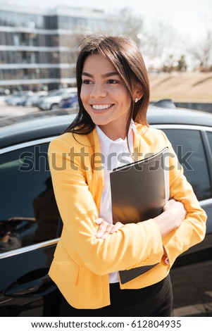 Happy beautiful young businesswoman holding folder standing with arms crossed near the car
