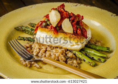 Fresh halibut fish fillet over bed farro with grilled asparagus, topped with salsa of red peppers, sundried tomatoes, garlic and capers sitting on rustic yellow plate with folk
