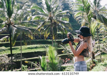 Young blonde hipster woman wearing black hat and jeans travelling around Asia, enjoying green rice terrace view, wanderlust blogger, Bali luxury vacation, taking pictures with her camera