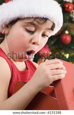 surprised girl wearing santa hat opening  a gift in front of a blurred decorated tree