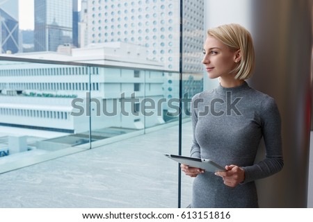 Thoughtful attractive blonde female administrative manager holding modern touchpad standing near window in coworking on urban background, charming pensive hipster girl admire cityscape from office Royalty-Free Stock Photo #613151816